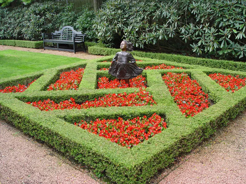 Statue of girl standing in breeze with red flowers and freshly trimmed hedges